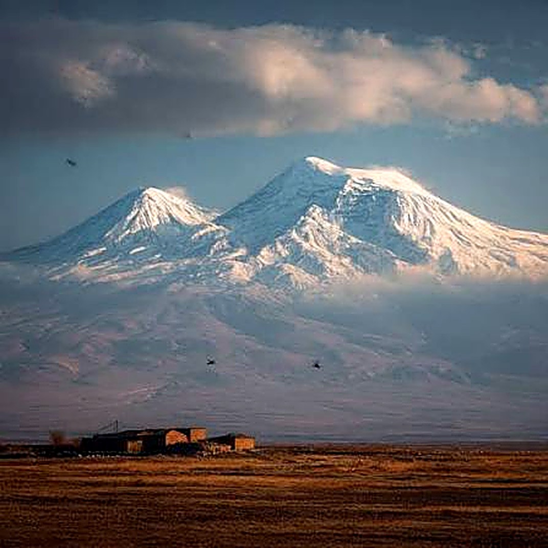 Masis and Sis, the twin peaks of Mount Ararat, rising over the Armenian plain at golden hour, with an abandoned farmstead in the foreground.